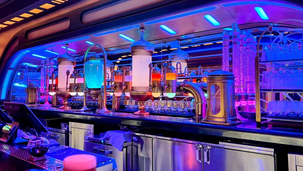 A futuristic bar counter illuminated by blue and purple lights, featuring unique drink dispensers with various colored liquids