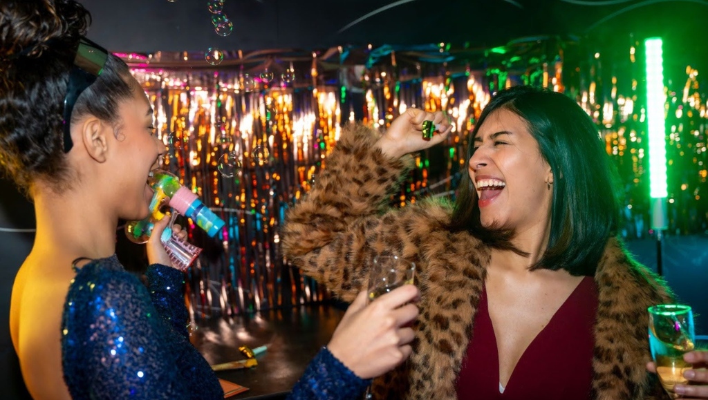 Two diverse young women laugh and dance at a vibrant party, one blowing bubbles with a toy gun while the other raises a glass of sparkling wine