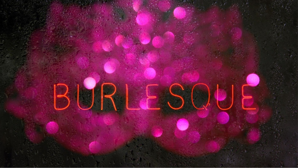 A vintage neon burlesque sign in a wet window with a hot pink sequined mask behind it